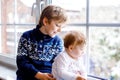 Happy adorable kid boy and cute baby girl sitting near window and looking outside on snow on Christmas day or morning Royalty Free Stock Photo