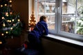 Happy adorable kid boy sitting near window and Christmas tree and looking outside on snow on Christmas day or morning Royalty Free Stock Photo