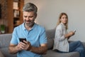 Happy adult caucasian husband with beard plays on smartphone, suffers from gadget addiction, ignores wife