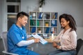 Happy adult chinese man doctor showing contract to young african american female patient in clinic