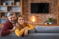 Happy adult european man and woman looking at camera, enjoy relaxing in living room interior with tv with blank screen