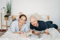 Happy adult married couple in casual clothing reading books on bed at bright interior