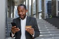 Happy african american businessman outside office building using phone and bank credit card for online shopping and Royalty Free Stock Photo