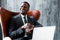 Happy african american businessman in a suit sitting on a chair and working at a laptop looking at the watch on his hand Royalty Free Stock Photo