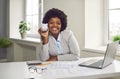 Happy African American businesswoman sitting at office desk, holding piggy bank and smiling Royalty Free Stock Photo