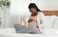 Happy Black Expectant Lady Enjoying Her Pregnancy, Sitting On Bed Touching Belly