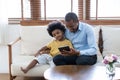 Happy African American father hugging his son, using digital tablet while sitting on sofa at home. Black family enjoying leisure, Royalty Free Stock Photo