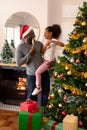 Happy african american father wearing santa hat and daughter decorating christmas tree