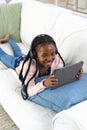 Happy african american girl lying on couch, listening to music and using tablet at home, copy space Royalty Free Stock Photo