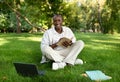 Happy african american guy studying with book and laptop in park, sitting in college campus and taking notes Royalty Free Stock Photo