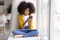 Happy african american little girl sitting on windowsill with smartphone Royalty Free Stock Photo