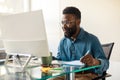 Happy african american man entrepreneur sitting at worktable, looking at computer screen and taking notes at office Royalty Free Stock Photo