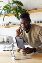 Happy african american man leaning on countertop in kitchen, using tablet and talking on smartphone Royalty Free Stock Photo