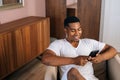 Happy African-American man sitting at armchair in cozy bright living room and using mobile phone. Royalty Free Stock Photo