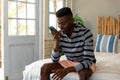 Happy african american man sitting on bed talking on smartphone at sunny holiday beach house Royalty Free Stock Photo