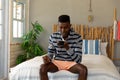 Happy african american man sitting on bed talking on smartphone at sunny holiday beach house Royalty Free Stock Photo