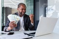 Happy african american man sitting in office at table in front of laptop, holding money cash in hands and enjoying Royalty Free Stock Photo