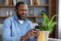 Happy african american man sitting on sofa at home and using mobile phone smiling. Close-up photo Royalty Free Stock Photo