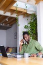 Happy african american man standing at table in kitchen, using laptop and talking on smartphone Royalty Free Stock Photo