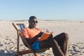 Happy african american man in sunglasses sitting in deckchair reading book on sunny beach Royalty Free Stock Photo