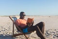 Happy african american man in sunglasses sitting in deckchair reading book on sunny beach Royalty Free Stock Photo