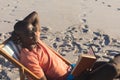 Happy african american man in sunglasses sitting in deckchair reading book on sunny beach Royalty Free Stock Photo