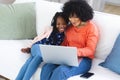 Happy african american mother and daughter sitting on couch and using laptop at home, copy space Royalty Free Stock Photo