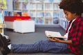 Happy african american schoolboy reading book sitting on floor in school library Royalty Free Stock Photo