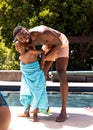 Happy african american shirtless father wrapped towel over son at poolside against trees in resort