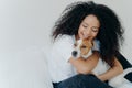 Happy African American woman expresses love to dog, embraces pet, dressed in casual wear, sit on comfortable bed in bedroom, Royalty Free Stock Photo