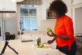 Happy african american woman in kitchen preparing health drink, making vlog using laptop and camera