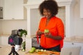 Happy african american woman in kitchen preparing health drink, making vlog using laptop and camera