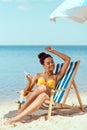 happy african american woman laying on deck chair and listening music in earphones with smartphone under beach umbrella in front