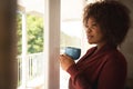 Happy african american woman looking out of sunny window, holding coffee cup and smiling Royalty Free Stock Photo