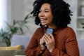 Happy African American woman sitting on sofa at home with phone in hands. She put her mobile phone to her chest and Royalty Free Stock Photo