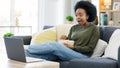 Happy african american woman streaming online movies on a laptop while snacking on popcorn and relaxing on a sofa at