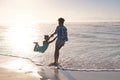 Happy african american young mother spinning playful daughter at sea shore against sky at sunset