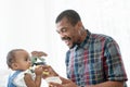 Happy African father feeding adorable toddler baby girl with spoon while sitting on sofa at home and kid`s face mess up with food