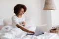 Happy african girl with laptop drinking cup of coffee, chatting with family and friends via video connection in bedroom Royalty Free Stock Photo