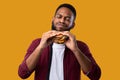 Happy African Man Eating Burger Posing Over Yellow Studio Background