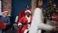 Happy african man in red santa claus costume sitting in the chair while smiling children jumping around him.