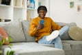 Happy african man using phone while sitting on sofa at his home. Concept of young people working on mobile devices. Royalty Free Stock Photo