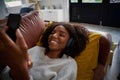 Happy african woman resting on couch using smartphone at home listening to music on headphones Royalty Free Stock Photo
