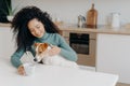 Happy Afro African woman with curly hairstyle treats dog in kitchen, pose at white table with mug of drink, enjoy domestic Royalty Free Stock Photo