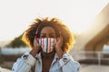 Happy Afro woman listening to playlist music with wireless headphones while wearing face colored mask Royalty Free Stock Photo