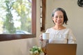 Happy aged Asian businesswoman remote working at the cafe, sipping coffee while using laptop Royalty Free Stock Photo