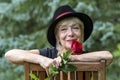 A happy aged woman, wearing a black hat, holding a gifted red rose against the background of nature.