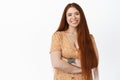 Happy ambitious girl with long red hair, smiling and laughing, cross arms on chest, posing in dress over white