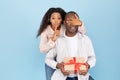 Happy anniversary. Portrait of young black woman covering her husband's eyes, making hush sigh, giving him gift box Royalty Free Stock Photo