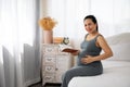 Happy Asia pregnant woman holding book sitting on white bed looking at camera Royalty Free Stock Photo